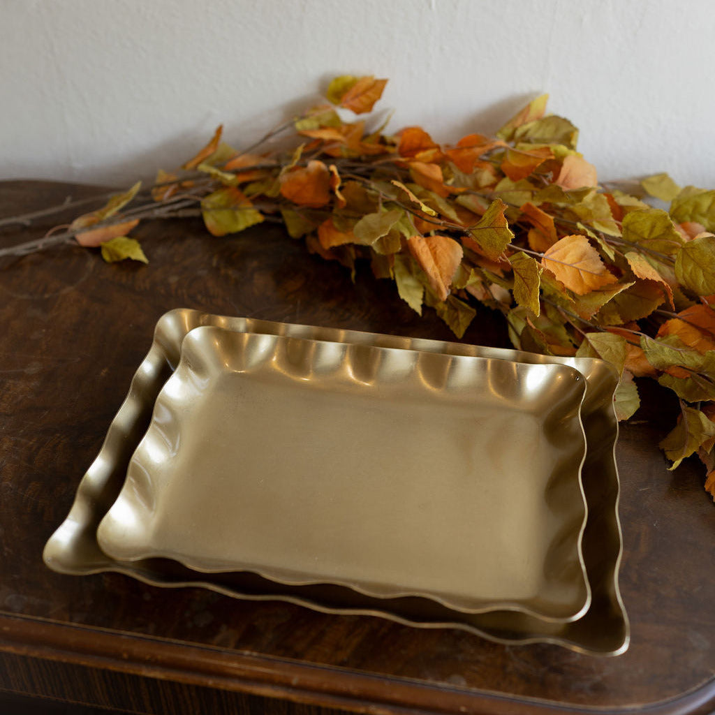 Two stacked rectangular serving trays with scalloped edges in a brushed gold finish, displayed on a dark wooden table with autumn leaves against a white wall.
