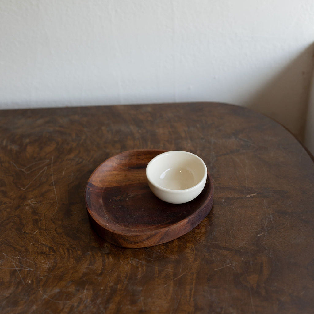 A small white ceramic bowl resting on a round dark wooden tray, which sits on a dark wooden table against a plain off-white wall.